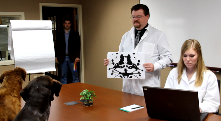 Two dogs sitting in a qualitative lab with pair of scientists.  The dog trainer is watching them on the other side of the room.